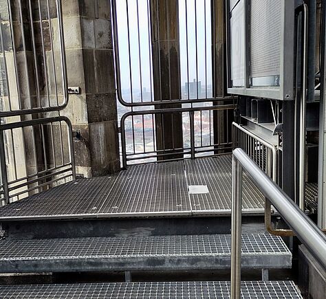 Mehrere Stufen aus grauem Metall mit offenem Gittermuster führen nach oben. Die Oberfläche ist gerastert und leicht rau, die Kanten sind klar und gerade, seitlich verlaufen glatte Metallgeländer.