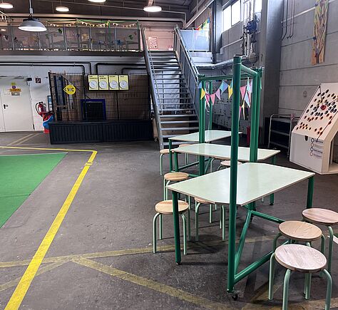 In einer geräumigen Halle mit hohen Decken stehen drei Tische, im Zickzack angeordnet, mit runden Hockern. Eine gelbe Linie markiert den Weg auf dem Boden zum WC. Im Hintergrund führt eine Metalltreppe mit Handläufen rechts und links und 18 Stufen zur Galerieebene. Die Halle ist hell ausgeleuchtet, Tageslicht fällt durch Fenster im oberen Bereich.