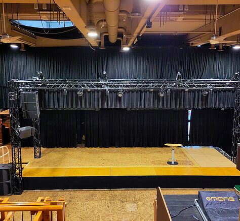 Die leere Bühne mit einem Holzboden und einem schwarzen Vorhang im Hintergrund. Über der Bühne, eine Konstruktion mit verschiedenen Scheinwerfern. Links und rechts Lautsprecher. Auf der Bühne ein einzelner Mikrofonständer in der Mitte.