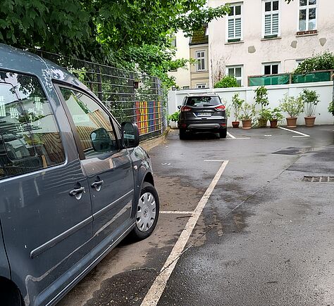 Asphaltierte Fläche, fast mittig im Bild ein weißer Streifen. Links neben diesem steht ein graues Auto, im Hinteren Bereich ein weiteres schwarzes. Zwischen den Auto ist ein Stück Zaun mit einer regenbogenfarbigen Verzierung zu sehen. Ganz hinten im Bild die Rückseite eine Wohnhauses. Davor eine halbhohe weiße Mauer, vor der einige Blumentöpfe stehen.