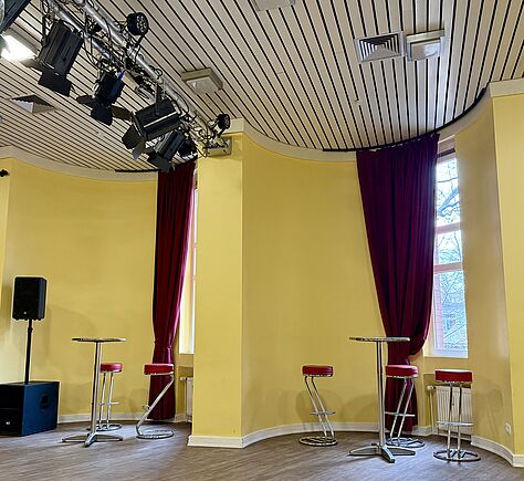 Die rechte Seite des Saales: gelbe Wänden, hohe Fenster in 2 halbrunden Erkern mit weinroten Vorhängen. In den Erkern steht jeweils ein Coktailtisch mit Barhockern mit roten Sitzflächen drumrum. Oben hängt eine Schiene mit vielen schwarzen Bühnenstrahlern. Links steht ein großer schwarzer Lautsprecher auf einem Stativ, darunter ein weiterer schwarzer Kastenlautsprecher.