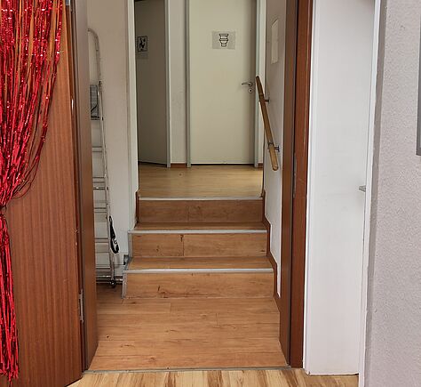 Blick durch eine offene Tür, die mit rotem Fransenvorhang behängt ist auf eine Treppe mit 3 hohen Stufen, rechts ein Handlauf. Der Boden ist durchgängig mit hellem Holz belegt, die Stufenkanten sind weiß markiert. Am Ende der Treppe ein Stück Flur quer dazu, 2 weiße Türen mit WC-Piktogrammen im Hintergrund. Rechts vor der Treppe ist ein breiter weißer Türrahmen zu sehen.