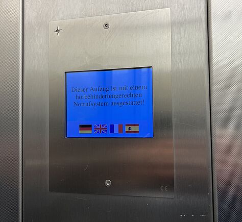Ein Schild mit dem Text: Dieser Fahrstuhl ist mit einem hörbehindertengerechten Notrufsystem ausgestattet. Darunter Flaggen; deutsch, englisch, französisch, spanisch.