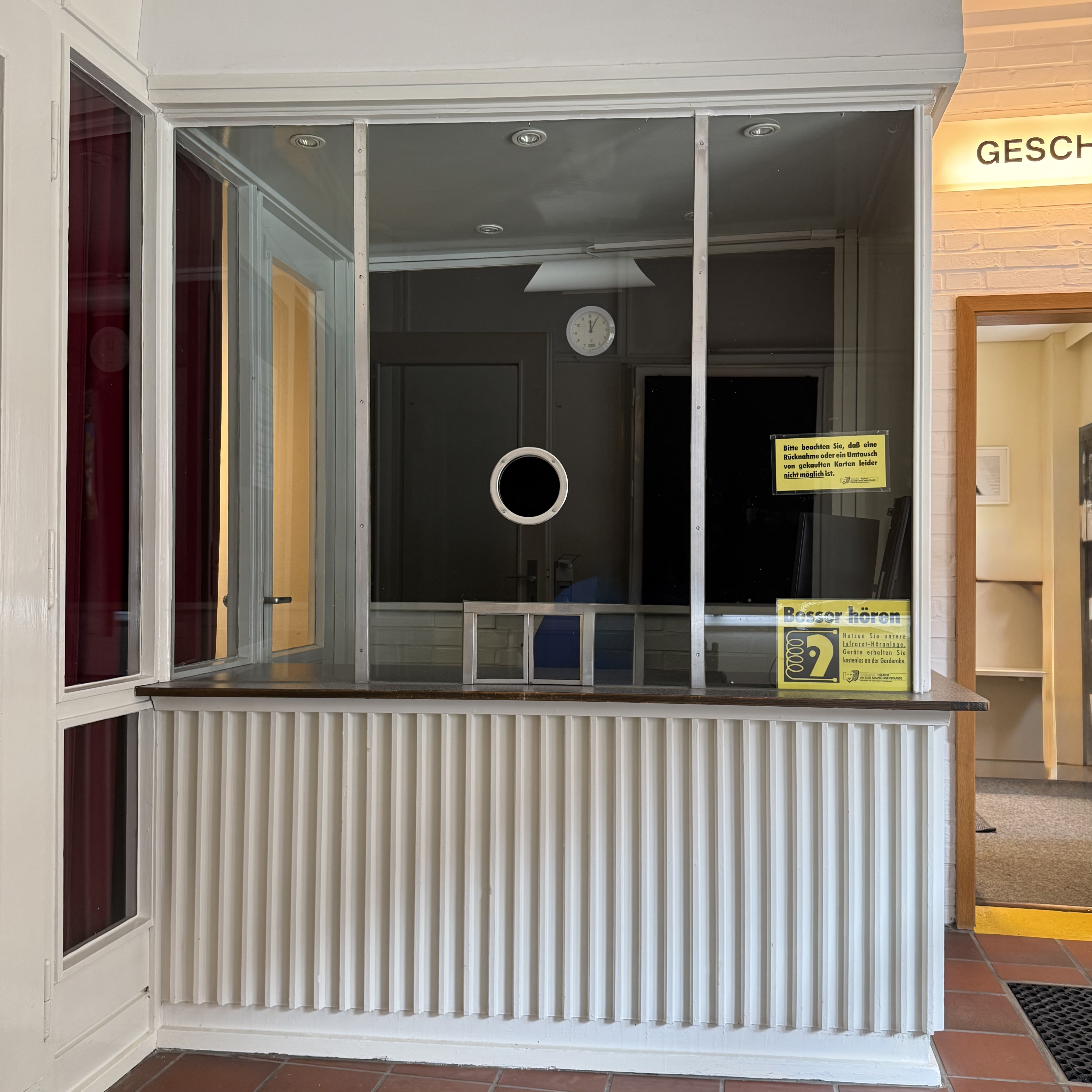 Über einer weißen Theke und einer dunklen Platte ist eine breite Glasscheibe mit Durchreiche und Sprechloch. Rechts davon führt eine Tür in das Gebäude.
