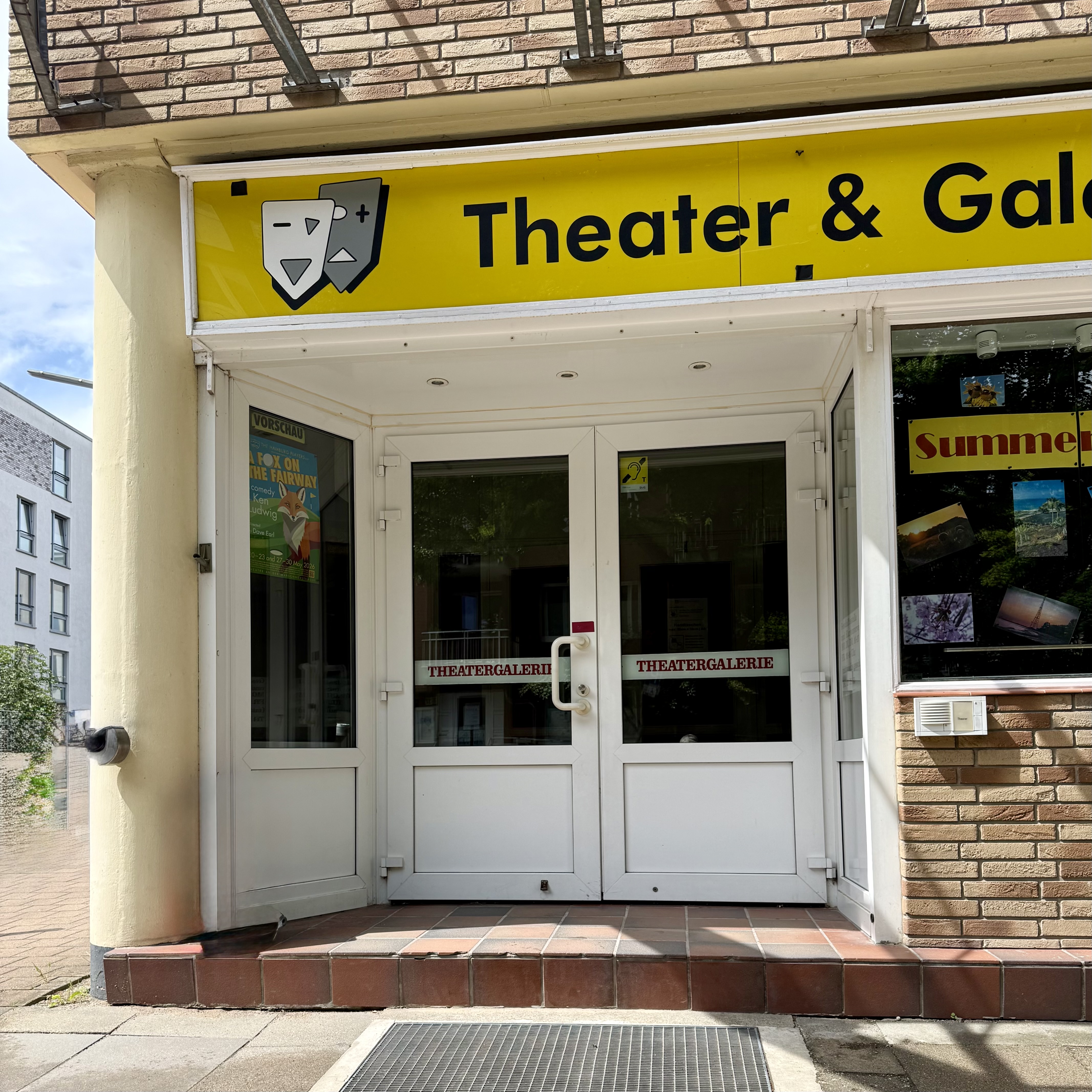 Auf dem Gehweg vor dem Eingang zum Theater ist ein Metallgitter. Es gibt eine Stufe. Die weiße Doppeltür mit Glaseinsätzen hat einen senkrechten Griff zum Öffnen. Eine Klingel mit Gegensprechanlage befindet sich rechts an der Hauswand unterhalb eines Fensters.