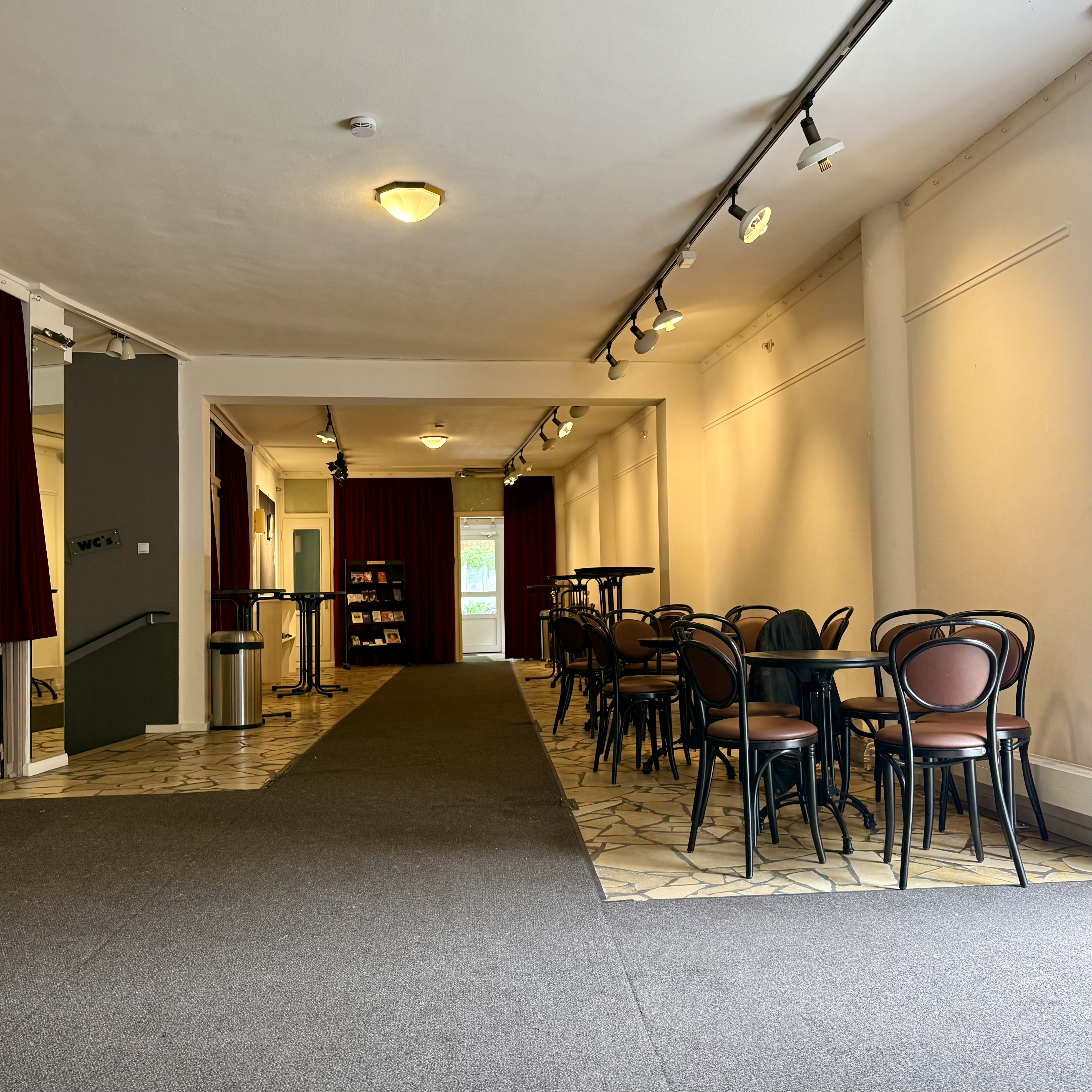 Das Foyer ist ein länglicher Raum mit hellem Wandanstrich und grauem Teppichboden in der Mitte und hellen unregelmäßigen Fliesen rechts und links. Vom Eingang kommend sind links ein paar Stehtische und mehrere runde Tische mit Stühlen. Rechts führt eine Treppe nach unten zum WC.