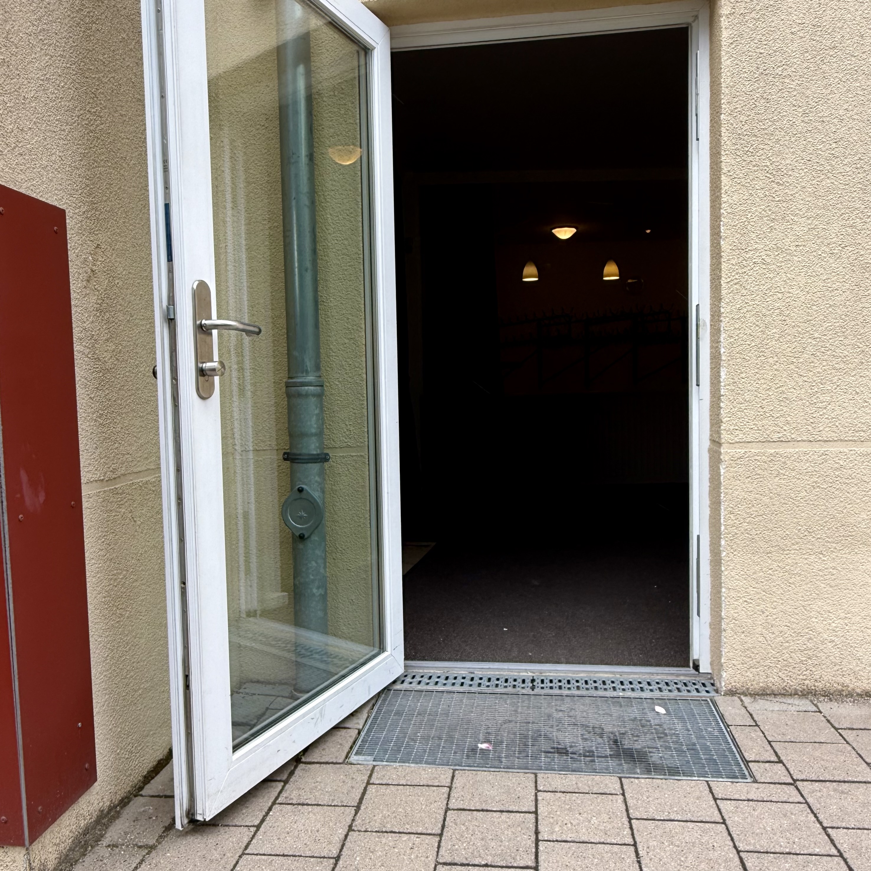 Eine offene Glastür mit weißem Rahmen führt ebenerdig in das Gebäude mit beigem Putz. Vor dem Eingang liegt ein Metallgitter. Links neben der Tür befindet sich ein rotes, rechteckiges Wandpaneel.