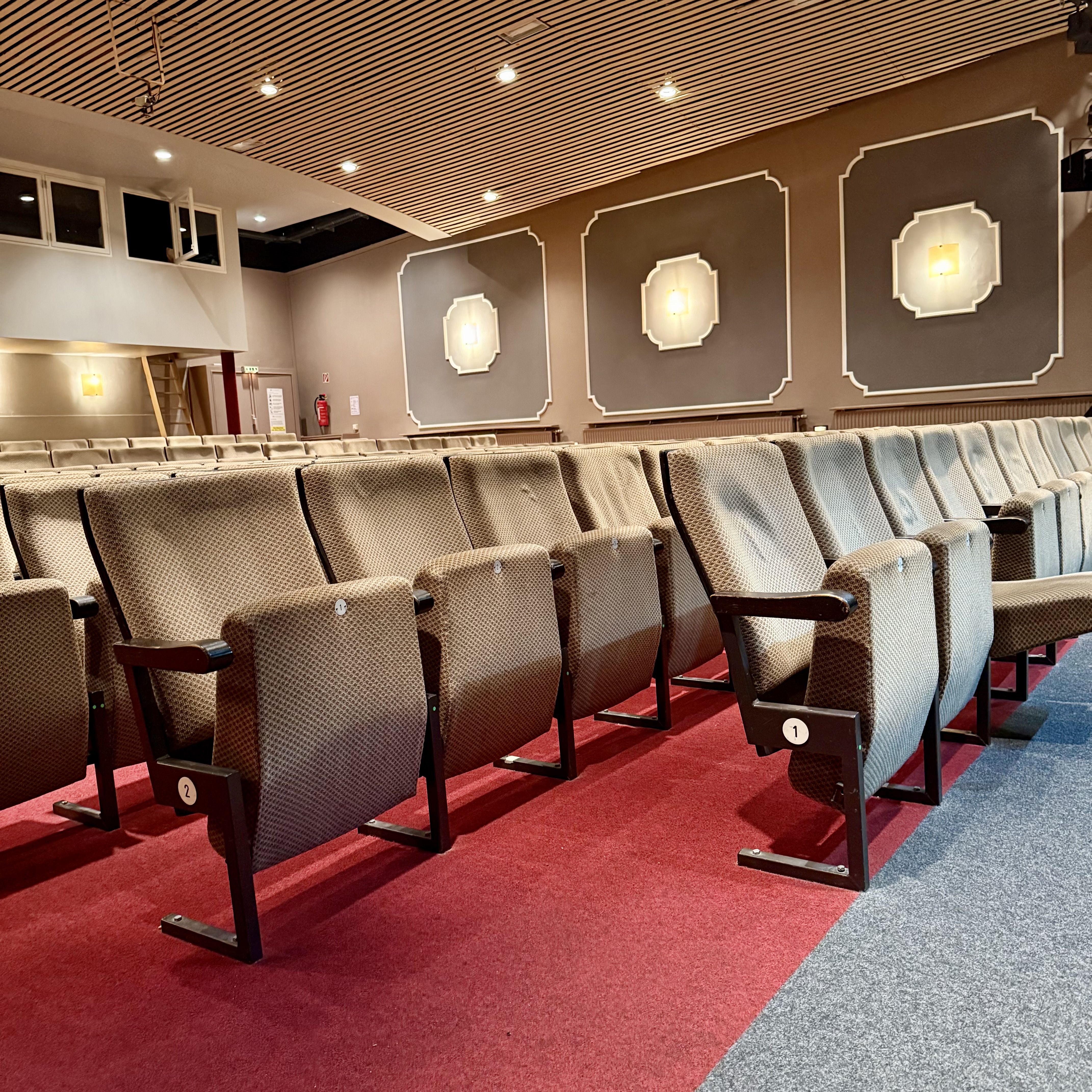 Im Theatersaal, die Klappsessel mit hellem Bezug stehen in mehreren Reihen. In der ersten Reihe ist Platz für ein oder zwei Rollstühle. Der Fußboden ist mit rotem und grauem Teppich belegt. Von den hellen Wänden und der Decke strahlen mehrere Leuchten.