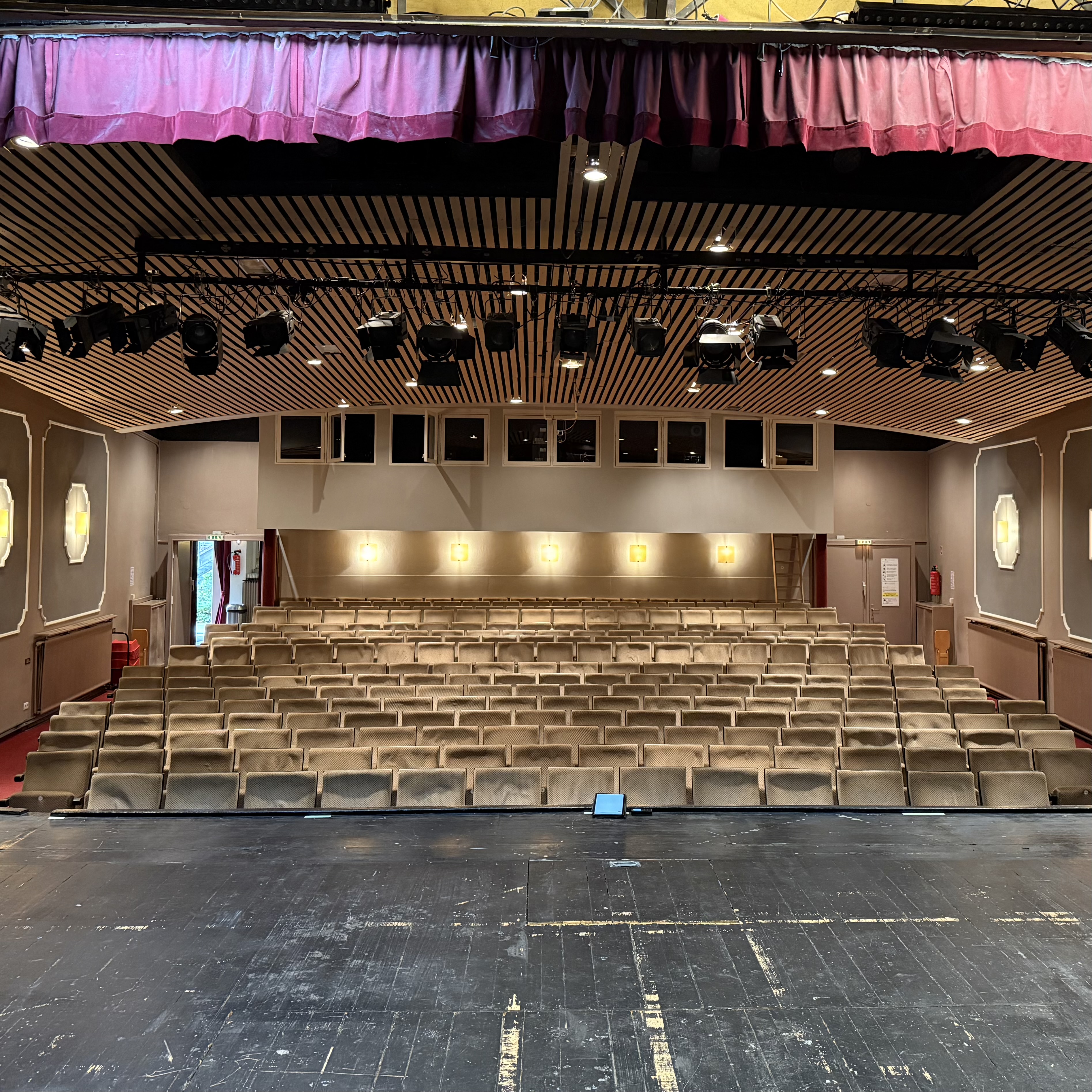 Der Saal mit den hellen Stuhlreihen ist von der Bühne aus fotografiert. An der Decke hängen zahlreiche Scheinwerfer. Über dem dunklen Bühnenboden hängt ein kurzer roter Vorhang.