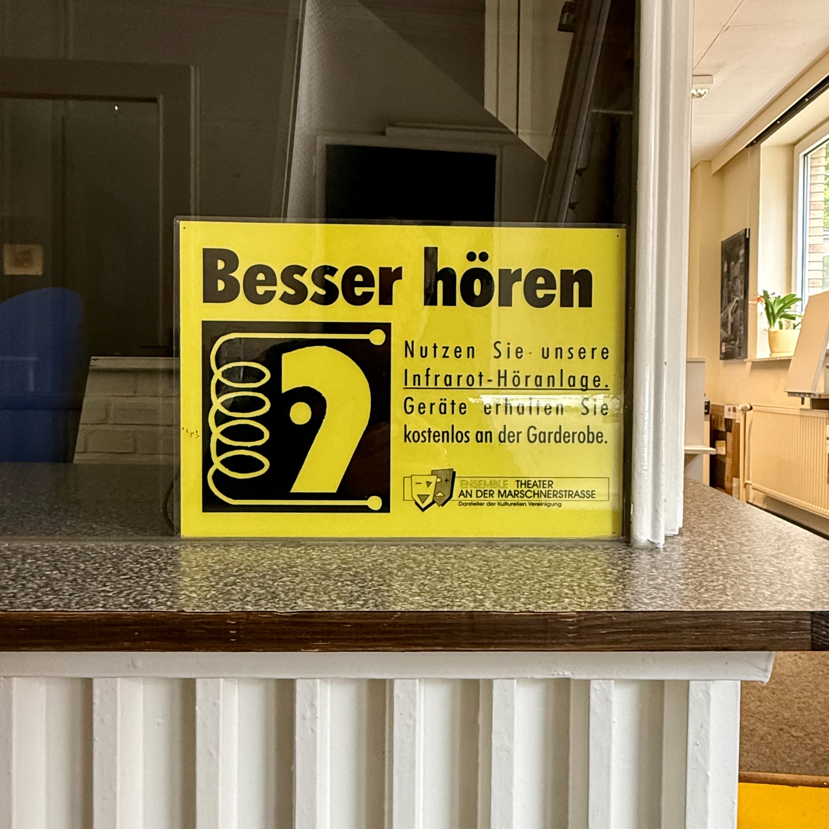 Ein gelbes Schild mit schwarzer Schrift weist auf die Möglichkeit hin, im Theater eine Infrarot-Höranlage zu nutzen. Geräte gibt es kostenlos an der Garderobe. Das Schild befindet sich an der Kasse.