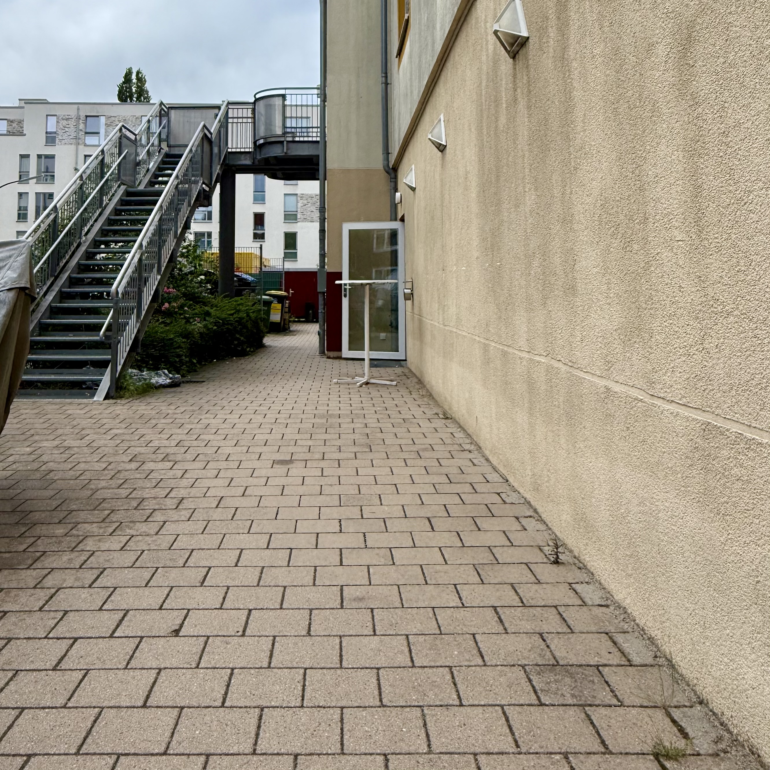 Ein gepflasterter Weg führt entlang einer beigen Gebäudewand zu dem Rollieingang rechts. Vor der Tür steht ein Stehtisch. Links ist eine lange Metalltreppe mit Geländer rechts und links und einem Absatz nach fünfzehn Stufen, die zu einer Plattform und einem Seiteneingang führt. Unterhalb der Treppe sind Büsche und Blumen.